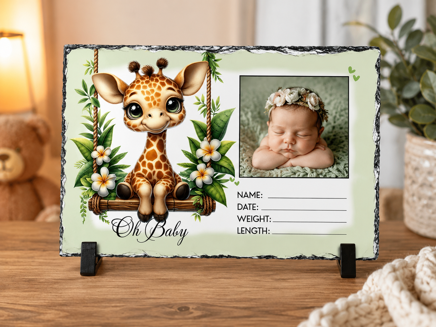 A personalized baby photo slate sign featuring a cute cartoon giraffe on a swing with the text 'Oh Baby' and a photo frame showing a sleeping newborn baby. The sign is displayed on a wooden table with a small black stand, with a warm, cozy background containing a teddy bear and plants.