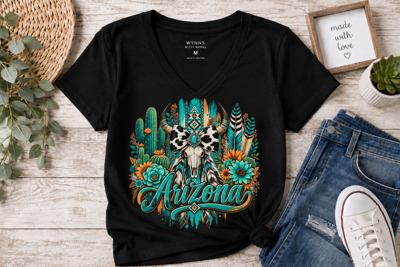 A black V-neck t-shirt with a colorful southwestern 'Arizona' graphic featuring a skull, cactus, and feathers, styled in a flat lay with jeans, a white sneaker, and a small framed sign on a light wood background.