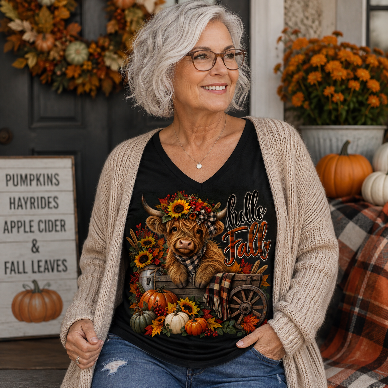 A smiling older woman with gray hair and glasses wears a black graphic t-shirt featuring a Highland cow in a wagon surrounded by autumn leaves and pumpkins, styled with a beige knit cardigan. She is posed in a cozy, decorated fall-themed porch setting with pumpkins, a sign, and orange flowers.