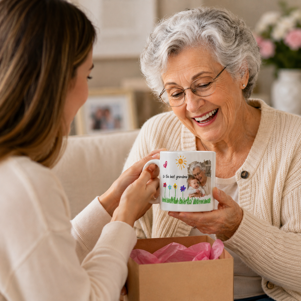 A smiling older woman holds a personalized white ceramic mug while a younger woman presents it. The mug features a printed photo of a cat and the text 'I love you grandma' with small colorful floral illustrations.