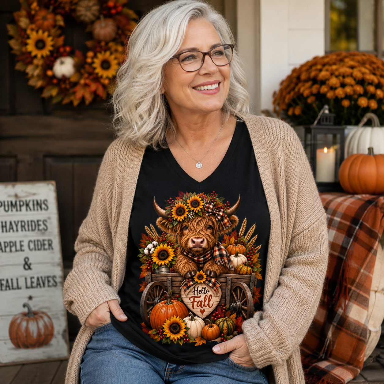 A smiling woman with gray hair and glasses wears a beige cardigan over a black graphic V-neck T-shirt featuring a Highland cow surrounded by sunflowers, pumpkins, and autumn leaves. She is posed on a decorated autumn porch with pumpkins, a wreath, and a plaid blanket in the background.