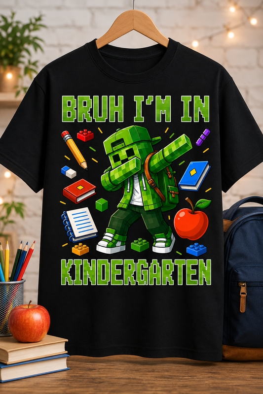 A black short-sleeve graphic T-shirt hanging on a wooden hanger against a softly blurred classroom-like background. The shirt features a green pixelated cartoon character in the center with the text 'BRUH I'M IN KINDERGARTEN' in large green letters, surrounded by school-themed items like a pencil, notebook, book, apple, and backpack.
