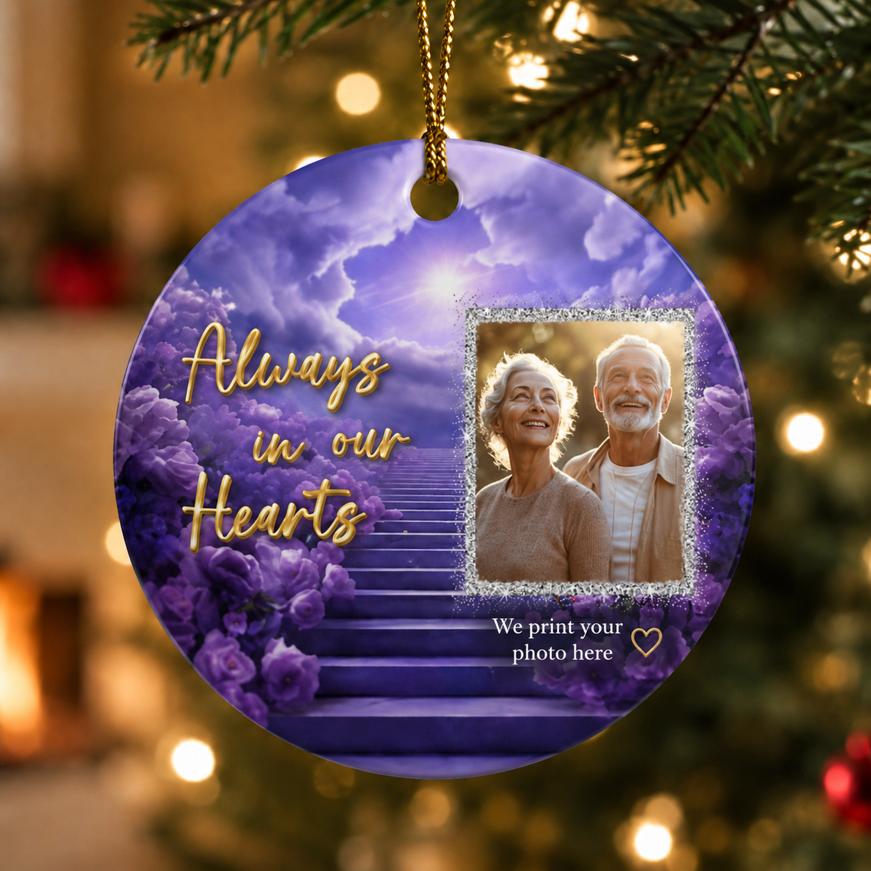 A round hanging ornament shaped like a Christmas tree branch, decorated with purple flowers, a staircase, and the words 'Always in our Hearts' in gold script. A sample photo of an elderly couple is printed in the center, with a gold hanging loop at the top.