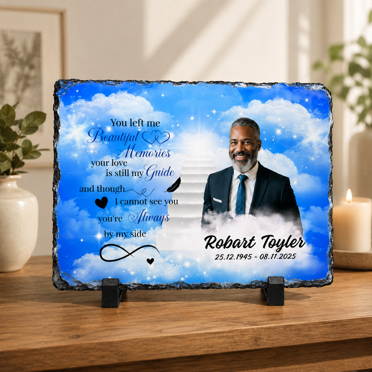 A personalized memorial slate plaque displayed on a wooden shelf. The slate features a printed blue sky and clouds design with a portrait of a man, memorial text, and the name 'Robart Toyler' with dates.