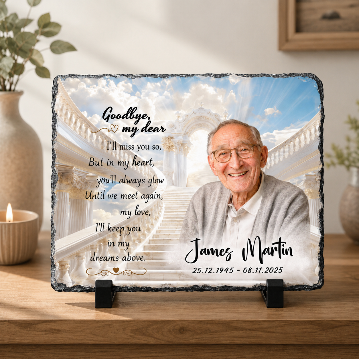 Personalized memorial slate plaque on a wooden table, featuring a photo of an elderly man, white staircase artwork, and printed remembrance text. The plaque is displayed on a stand in a softly lit indoor setting with plants and a candle in the background.