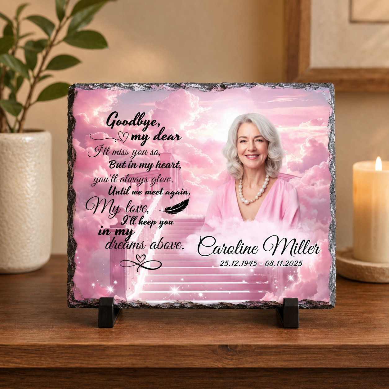 Personalized memorial slate plaque on a wooden stand, featuring a pink sky and clouds background, a portrait of an elderly woman, and printed remembrance text for Caroline Miller, styled on a wooden table with a lit candle and potted plant in the background.