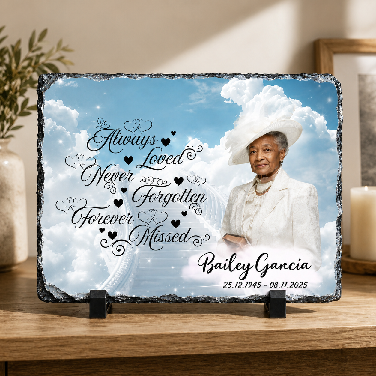 Personalized memorial slate plaque on a wooden stand, featuring a photo of an elderly woman in a white outfit against a blue sky and clouds, with the words 'Always Loved Never Forgotten Forever Missed' and the name 'Bailey Garcia' plus dates, displayed on a tabletop in a home setting.