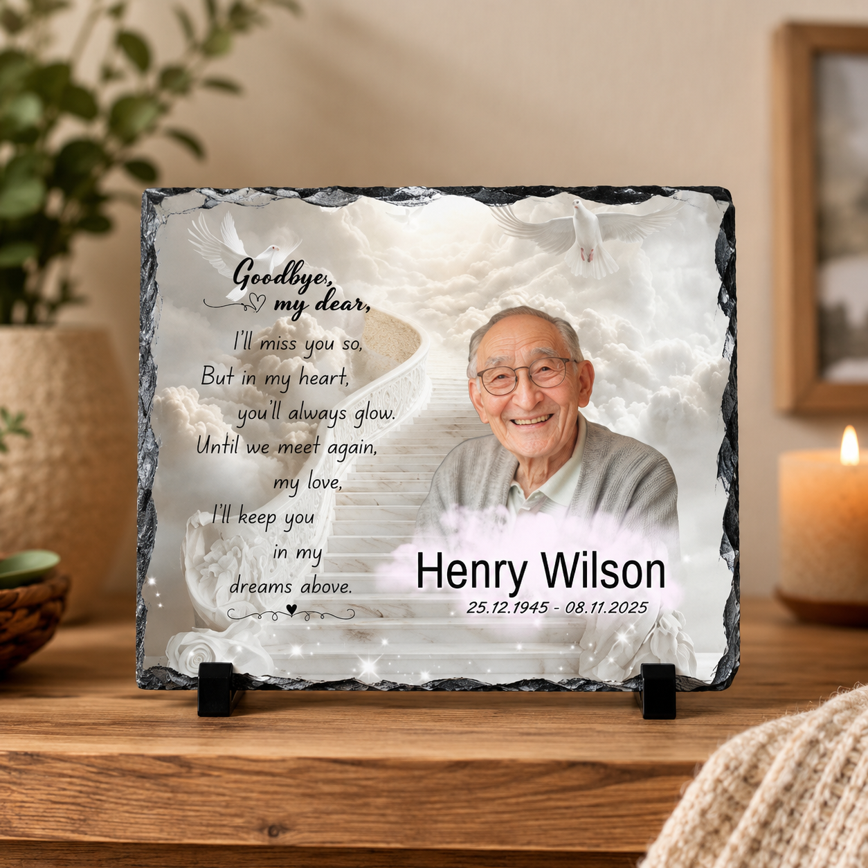 Personalized memorial slate plaque on a wooden table, featuring a printed portrait of an elderly man, white doves, a poem, and the name 'Henry Wilson' with dates. The plaque is displayed in a warm, home-like setting with a candle and framed picture in the background.