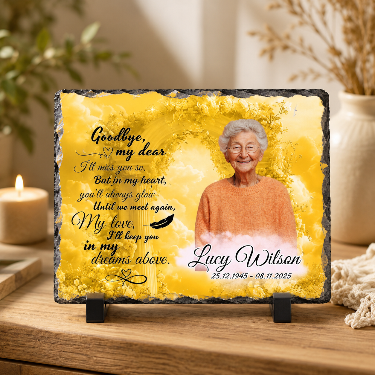 Personalized memorial slate plaque on a wooden table with a candle and dried flowers in the background. The plaque has a yellow watercolor design with a portrait of an elderly woman, black text reading 'Goodbye, my dear.
