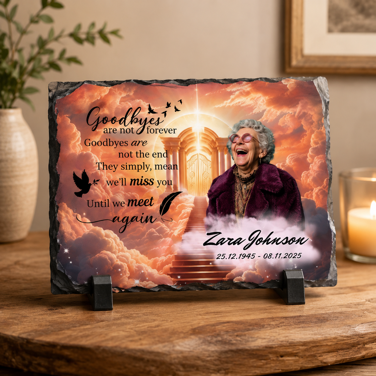 Personalized memorial slate plaque on a wooden table, featuring a smiling elderly woman in a purple coat against a glowing clouds-and-stairs background, with the text 'Goodbyes are not forever' and the name 'Zara Johnson' plus dates. A candle, vase, and framed art are softly visible in the room.