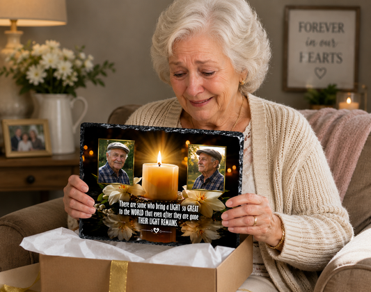 An elderly woman with white hair sits on a couch in a cozy living room, holding a printed memorial photo frame over an open gift box. The frame features a candle, flowers, and a quote, while the woman looks down with a tearful expression.