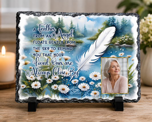 Personalized memorial slate plaque with a scenic landscape print, a large white feather graphic, blue cursive text, white daisies, and a small inset photo of a woman, displayed on a wooden stand.