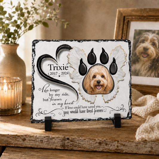 Personalized pet memorial slate plaque on a rustic wooden table, featuring a photo of a golden doodle dog inside an angel wing and heart-shaped paw print design, with a candle and framed photo in the background.
