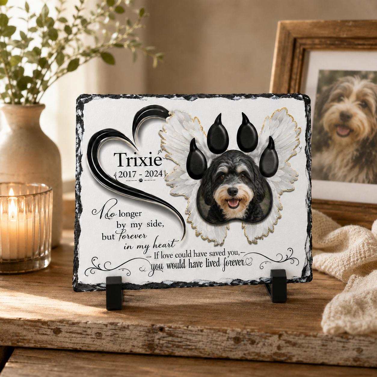 A memorial slate plaque displayed on a rustic wooden table, featuring a black and white dog portrait with angel wings, a large paw print, and the name 'Trixie' with dates and remembrance text. The scene is styled with a lit candle, a vase, and a framed picture in the background.
