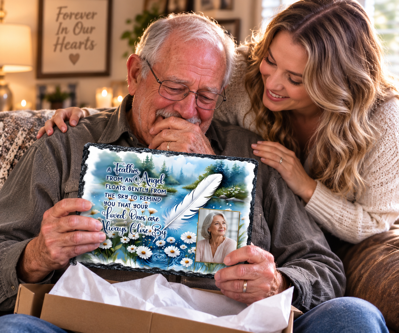 An elderly man and a woman are sitting together on a couch, looking at a custom pet memorial plaque held up from an open box. The plaque features a blue and white landscape with a white feather, daisies, a photo of the pet, and a sentimental quote.