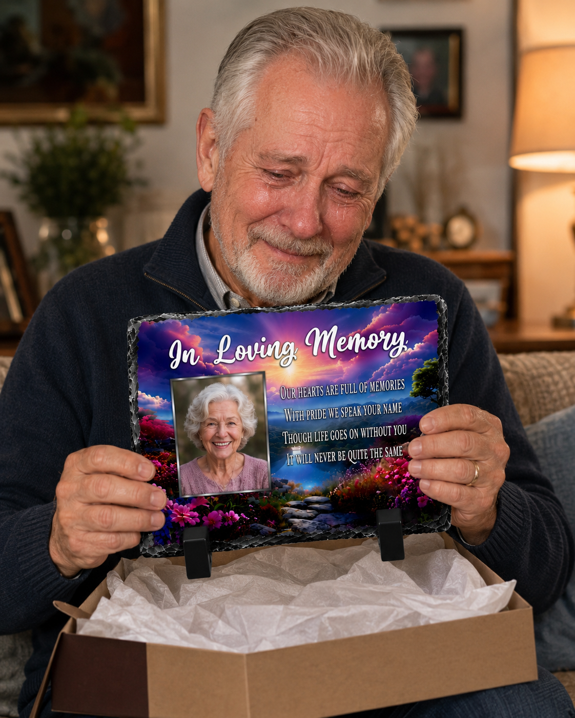 An older man sitting in a cozy living room setting holds up a personalized memorial slate plaque. The plaque features a colorful sunset landscape, a photo of the man, and the text 'In Loving Memory' along with a memorial message.