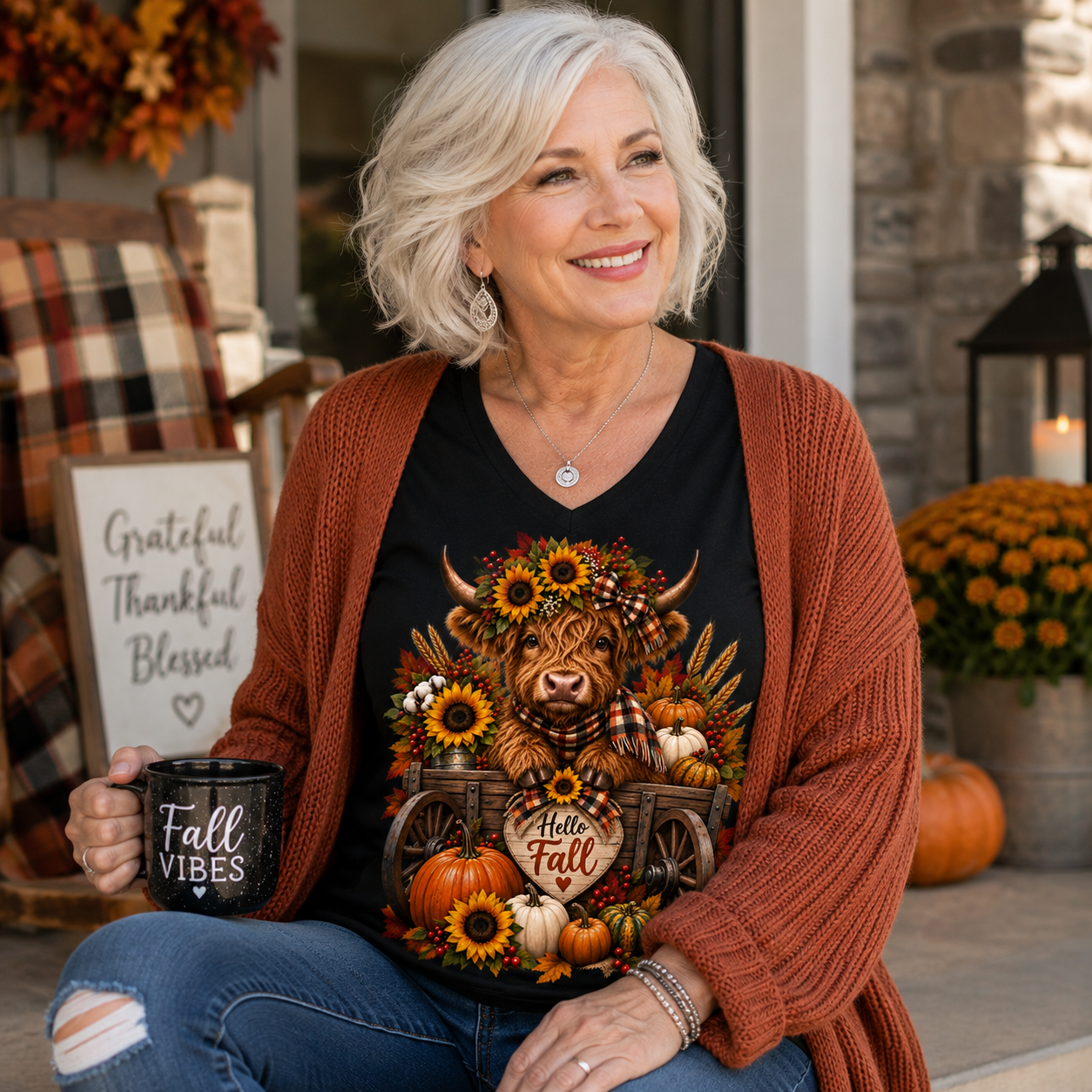 A smiling woman with short gray hair sits on a porch wearing a black graphic T-shirt with a fall-themed cow illustration and a rust-brown cardigan. She holds a black mug that says 'Fall Vibes' and wears blue jeans.