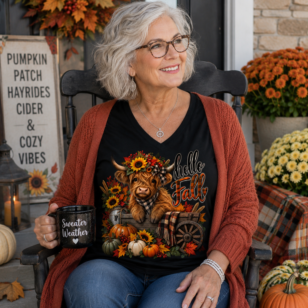 An older woman with gray hair and glasses sits in a rocking chair wearing a black graphic t-shirt with a Highland cow and autumn design, a rust-colored cardigan, and blue jeans. She holds a black mug that reads 'Sweater Weather'.