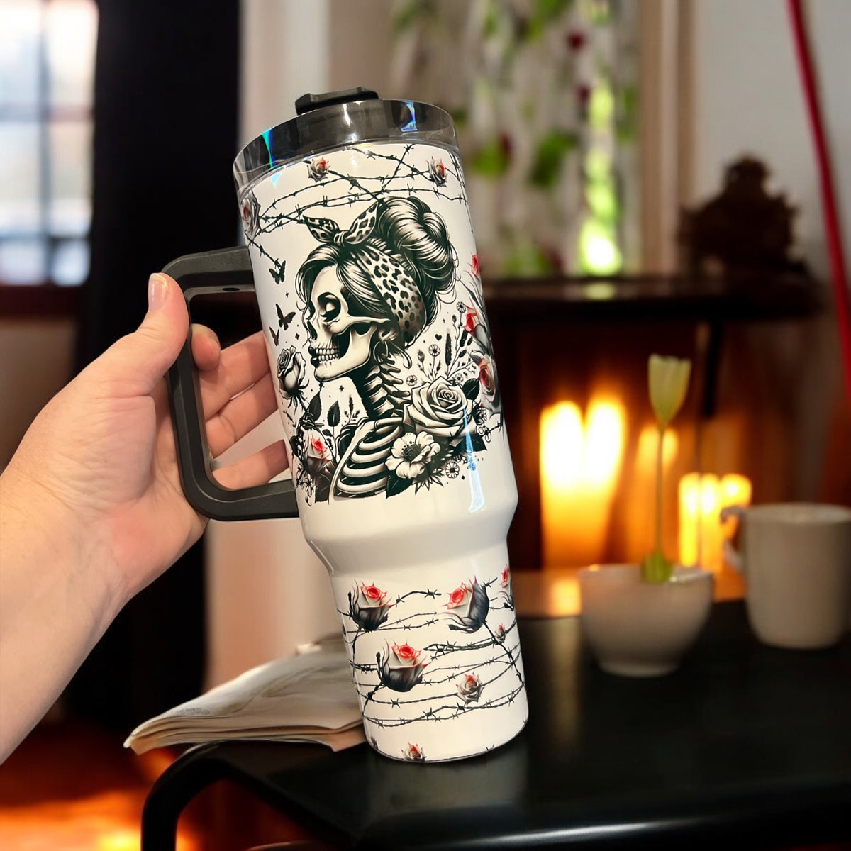 Person holding a white tumbler with black skull and floral design in a cozy room.