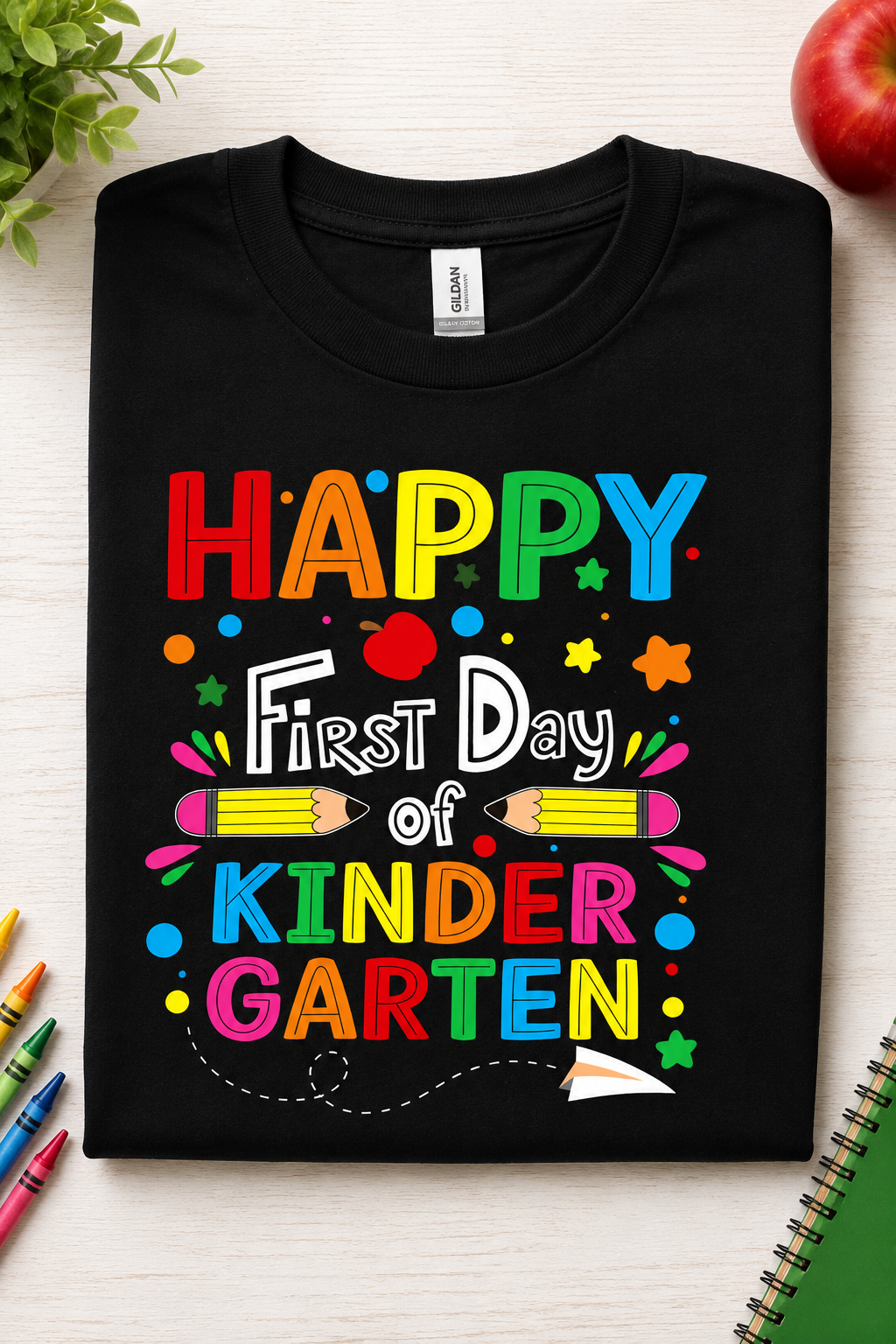 A folded black t-shirt with a colorful 'Happy First Day of Kinder Garten' graphic print, displayed on a light wood background with school-themed props like crayons, a red apple, and a notebook.