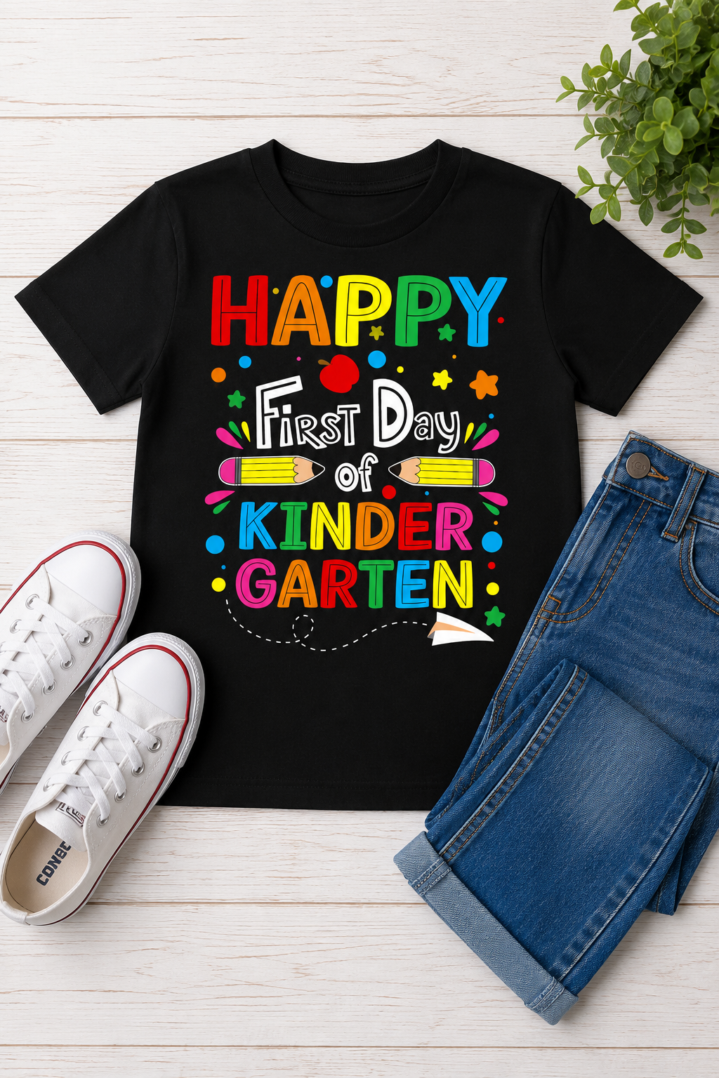 Black Happy First Day of Kindergarten kids shirt displayed in a bright back-to-school flat lay with jeans, sneakers, an apple, and colorful school supplies.