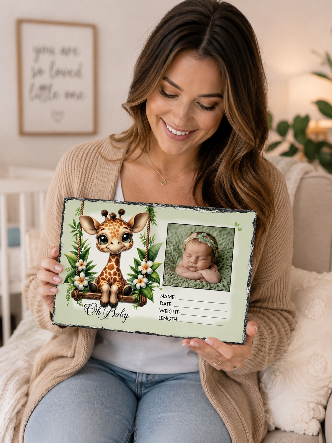 A smiling woman sits on a couch holding a personalized baby keepsake photo board. The board has a light green background with a cute cartoon giraffe illustration, the text 'Oh Baby,' and a photo slot for a newborn baby picture.