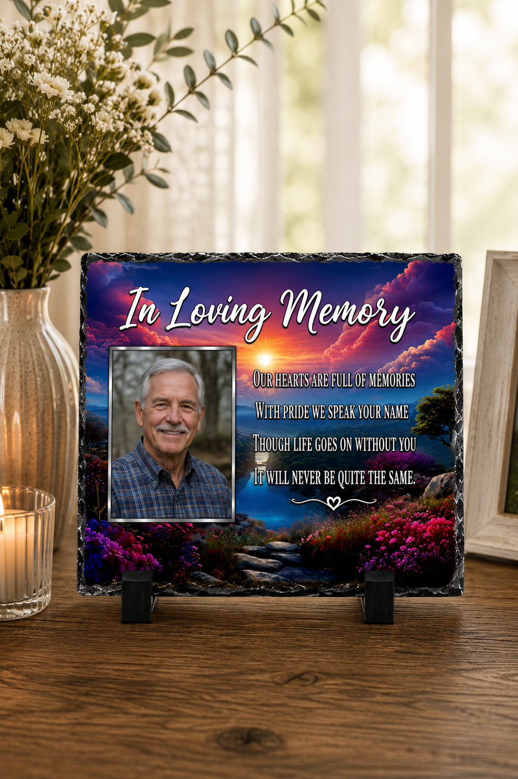 A personalized memorial slate plaque displayed on a wooden table, featuring a photo of a man, the text 'In Loving Memory,' and a scenic sunset landscape with flowers. The plaque is styled in a home setting with a vase of white flowers and a candle in the background.