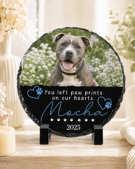 A round black slate memorial plaque on a small black stand, featuring a photo of a gray and white pitbull-type dog in a field of white flowers, with the text 'You left paw prints on our hearts. Mocha 2025' on a black section.