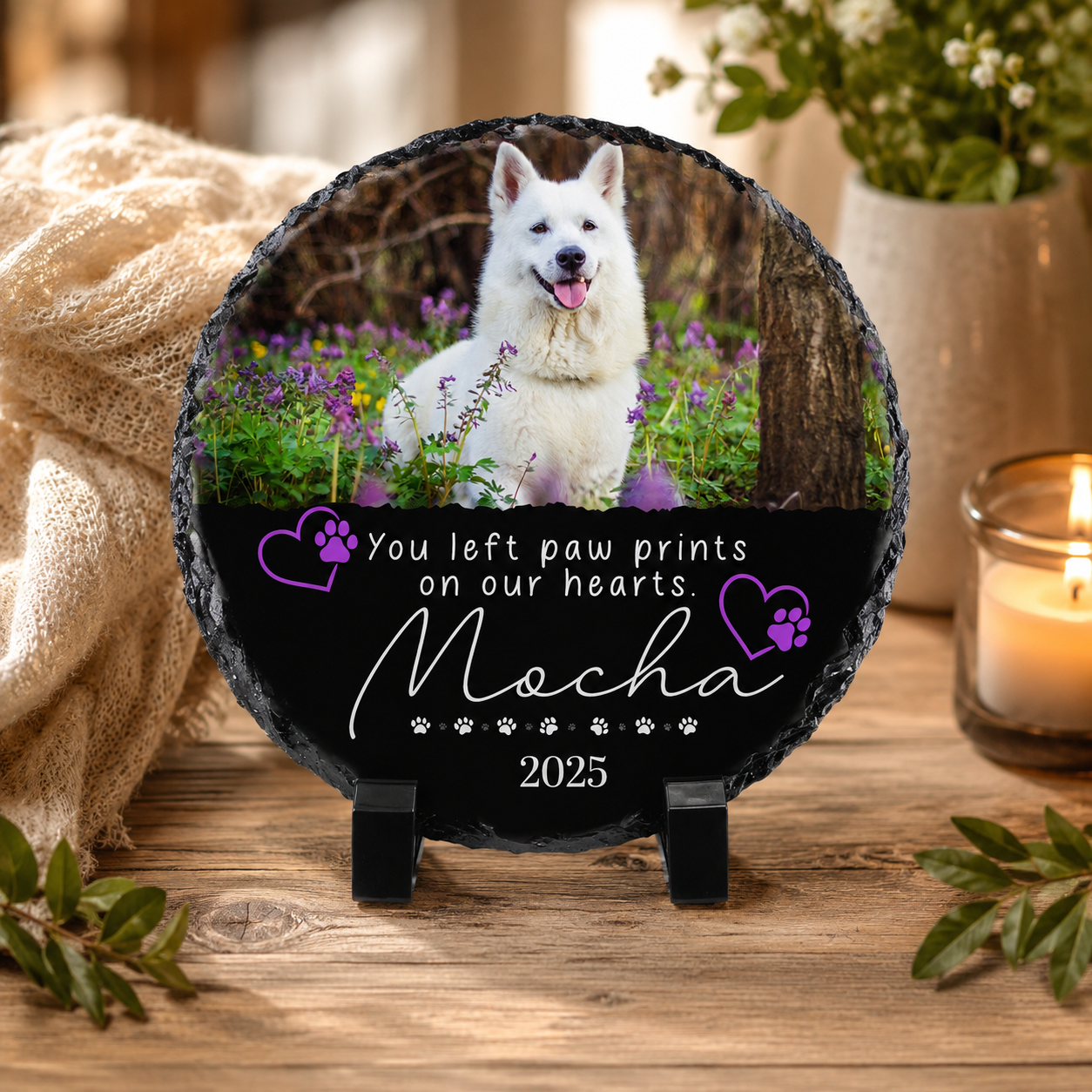 Personalized pet memorial slate plaque on a wooden table, featuring a photo of a white dog, purple paw print hearts, and the text 'You left paw prints on our hearts. Mocha 2025'.