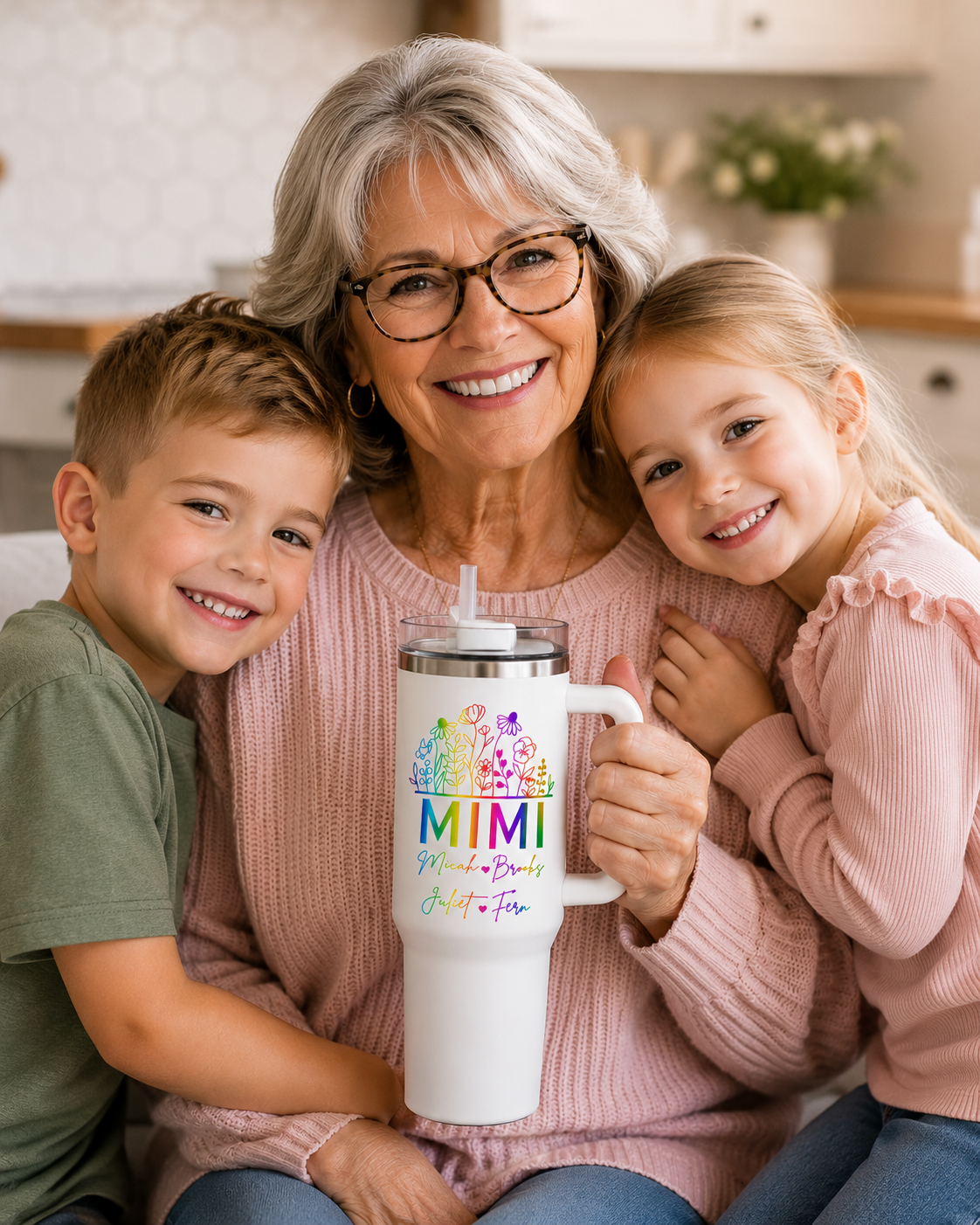 A smiling older woman with gray hair and glasses is seated between two children in a bright indoor setting. She holds a tall white insulated tumbler with a clear lid and a colorful printed design that reads 'MIMI' with smaller text below.