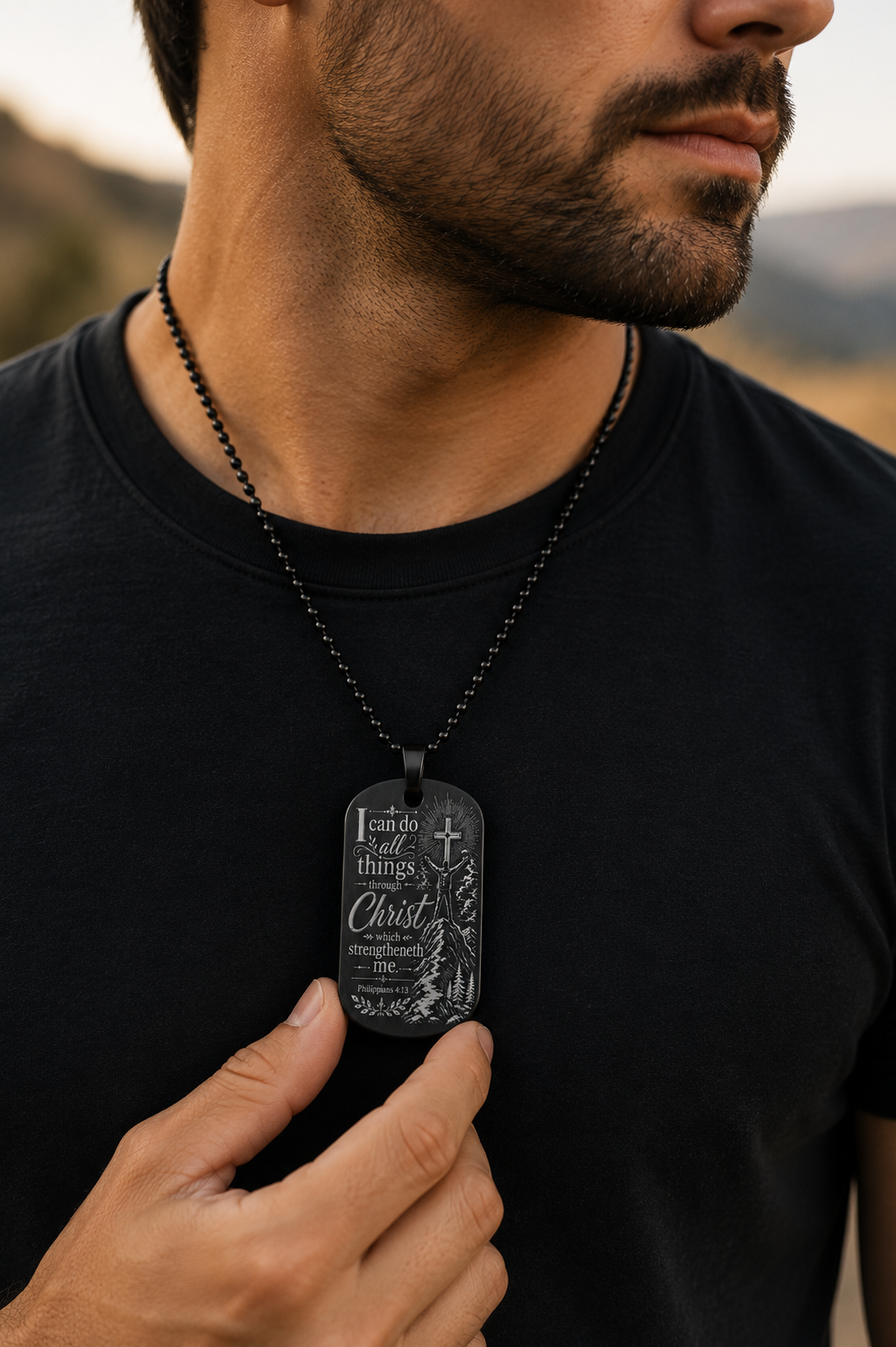 Close-up of a man wearing a black Philippians 4:13 engraved dog tag necklace featuring a cross and mountain design. Inspirational Christian jewelry reminding believers, "I can do all things through Christ who strengthens me."